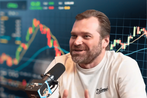 DraftKings co-founder Matt Kalish speaking into microphone discussing prediction markets with stock trading charts behind him during sportsbook industry interview. DraftKings cofounder blasts Kalshi prediction markets amid growing sportsbook industry tensions