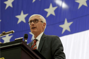 Wisconsin Governor Evers speaking at podium with American flag backdrop addressing tribal mobile sports betting legislation. Gov. Evers bars Wisconsin employees from exploiting insider information through prediction markets
