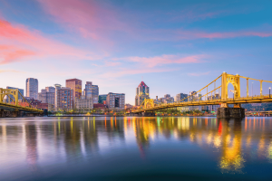 Pittsburgh skyline at sunset with illuminated yellow bridges reflecting across the Allegheny River in downtown Pennsylvania. Pennsylvania regulators approve $180K in gaming fines while expanding involuntary exclusion lists