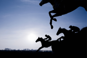 Silhouetted jockeys ride racehorses over a steeplechase jump during a horse racing event, illustrating thoroughbred racing and equestrian sport against a dramatic sky. Hawthorne creditors seek probe into racing funds amid bankruptcy dispute case