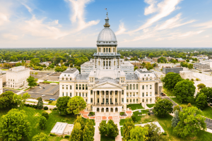 Aerial view of the Illinois State Capitol building in Springfield, Illinois, showcasing the historic government landmark, iconic dome, landscaped grounds, and downtown skyline under a bright blue sky. Illinois budget bill targets sports prediction markets with unique transaction tax