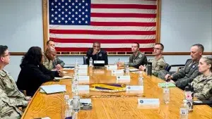Don Davis seated at a conference table with military personnel in front of a large American flag.