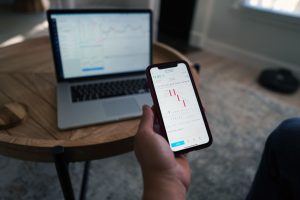 Person holding a smartphone with a trading chart in front of a laptop showing market data